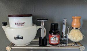 Shaving Kit Dec 2016 - a white shaving mug with a tub resting inside with the label Tabac on it. S silver safety razor stands vertically in a short black holder. There is a small brown bottle in the centre labelled Badger pre-shave oil. To the right is another silver safety razor, and on the far right is a wooden-handled shaving brush resting on its stand.