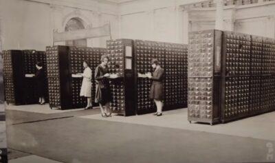 Rows of old wooden filing cabinets with tiny drawers, and some women cataloguers