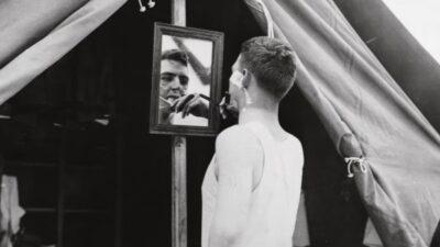 Black and white photo of a man shaving with a cut throat razor, whilst looking into a mirror that is mounted on a wooden pole holding up a tent