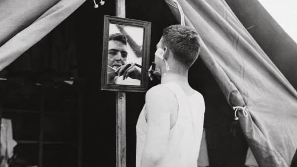Black and white photo of a man shaving with a cut throat razor, whilst looking into a mirror that is mounted on a wooden pole holding up a tent
