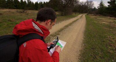 A man in a red jacket, holding a GPS device and a map, standing on a gravel road stretching out to the distance