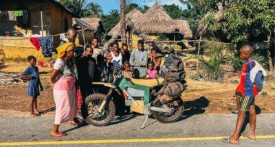 CAKE offroad motorcycle surrounded by onlookers in Africa, with thatch covered houses in background