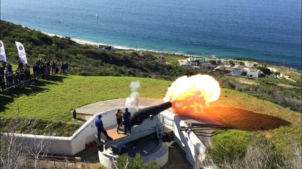 Foreground shows an old cannon being being fired with smoke and flame coming out of the barrel, with some grass hill behind it leading down to the sea in the background