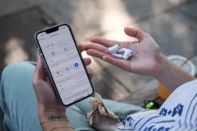 Smartphone held in the left hand with a pair of white ear buds in the palm of the right hand.