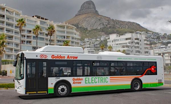 A whte commuter bus with words Golden Arrow and Electric on the side, with the background showing 8-story flats and in the far distance a hill peak.