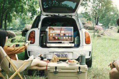 Two people sitting in a grassy picnic area, one with feet on a coller box, looking at a TV that is standing in the rear door of a mini hatchback car.