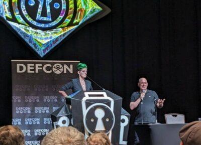 A stage with sign in background showing DEFCON, and a lady with green coloured hair standing behind a podium, with a bald-looking man standing beside here holding a microphone in his right hand.
