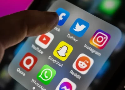 Phone screen, with a folder open showing icons for apps such as Twitter, Facebook, Instagram, YouTube, Snapchat, Reddit, etc. An index finger is hovering over the Facebook icon.