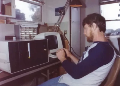 Man sitting at a desk, typing on a retro computer keyboard with a small integrated display. On the left stands a larger rectangular cabinet with a floppy disc protruding from it.