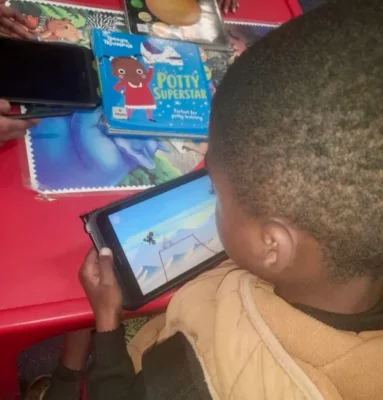 Child holding a tablet, with a game being played on the screen. In the background is a red plastic table with various books on it.