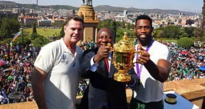 Two SA rugby players holding up the Rugby World Cup tother with the South African President, with a big crowd in the background