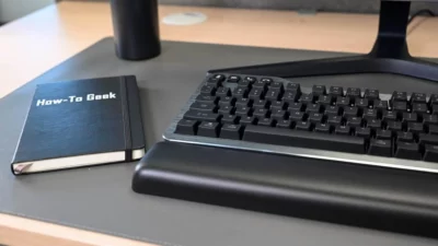 Half of a keyboard showing on a desk, with a black covered journal next to. Title on cover of the journal says How-To Geek.