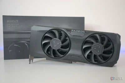 Black coloured Graphics Processor Unit with two fans on the front and the name RADEON on it. Behind it is a black box with words AMD Radeon RX7900XT on it.