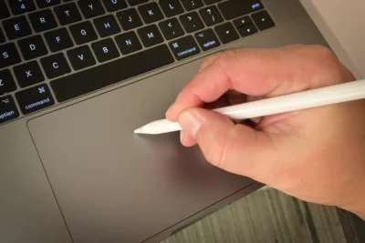 Close-up partial view of a Macbook keyboard, and showing a hand holding a stylus, poised over the trackpad for writing.