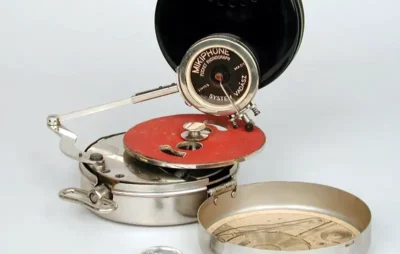 Silver tin with top open and a wind-up handle on the side. On top, and offset slightly, is an orange flat disc. Above that is a vertical disc with words Mikrophone pocket phonograph on the side, with a needle protruding from the bottom down to the orange disc.