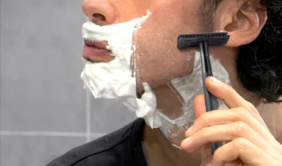 Close side-view of a man's cheek partially covered in shaving soap, and his hand holding a matt-black safety razor against his cheek.