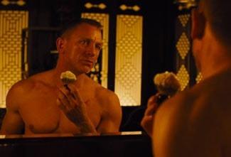 Daniel Craig, as James Bond, standing in front of a mirror and lathering his face with a shaving brush with shaving cream on it