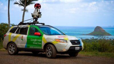 A car with Google street map green and while colours, with a camera mounted on the roof. Behind it in the distance the sea can be seen.