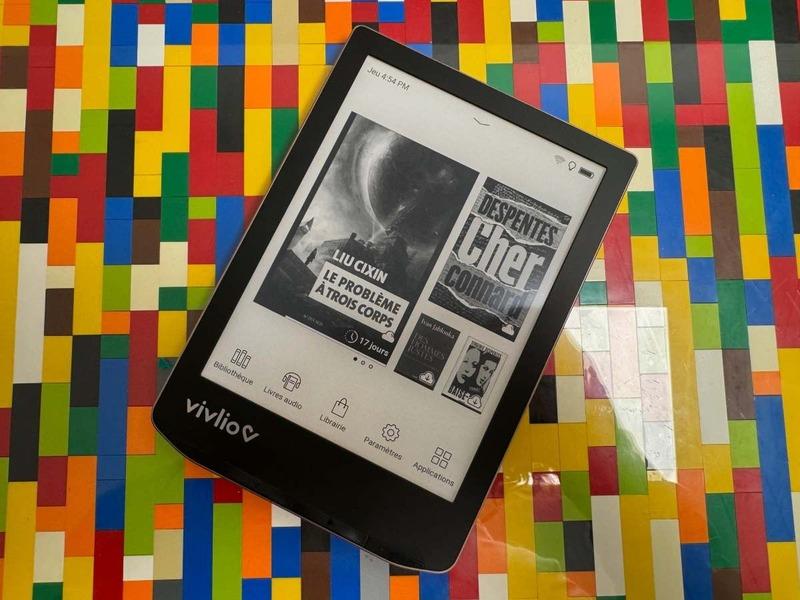An e-book reader lying on a multi-coloured background. The reader shows some black and white book titles in tiles on its front page.