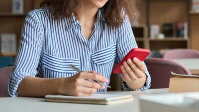 Woman sitting at a desk holding a pen in her right hand which is hovering over a paper notebook on the desk, and her left hand is holding a red phone