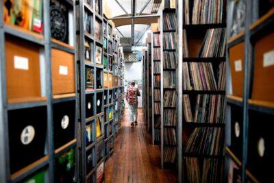 Tall library shelfves that show vinyl records stacked on the shelves. A man is seen walking between the rows of shelves.