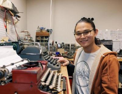 Young man smiling and sitting at desk with two typewriters on the desk in front of him. Clsoest one is a red colour and the one behind is an olive blue colour. The far wall in the background shows drawers and tools stacked up like a workbench.