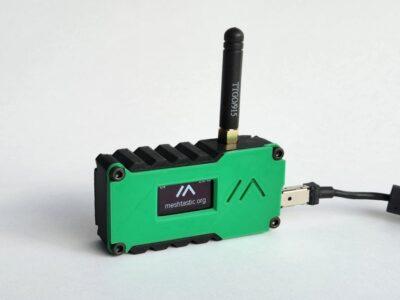 A green and black plastic box that is a little larger than a thumb. It has a small OLED display on the front saying "meshtastic.org". Protruding from the top is a short stubby black antenna, and on the right is a USB power cable.