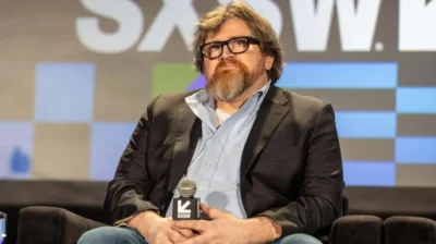 Bearded man with glasses sitting ona couch holdinga microphone in his hands. Behind him looks like a display you'd see as a backdrop on a stage.