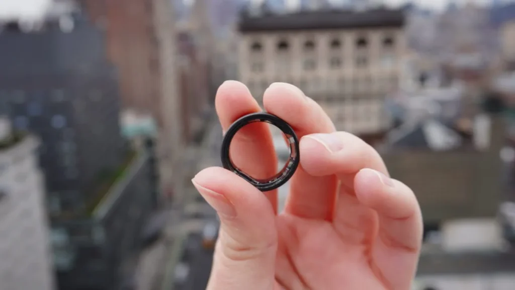 Fingers of a hand, holding a dark coloured ring. In the background is a blurry view of a city.