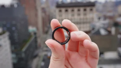 Fingers of a hand, holding a dark coloured ring. In the background is a blurry view of a city.