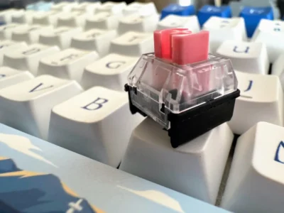 Close up view of a keyboard showing just a few white keys, with a cube shaped key switch resting on top of one of the keys. The switch is made of plastic with a black coloured base, and a transparent top. Protruding from the top of the switch is a few mm's of pink stalk that the key cap would be attached to.