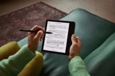 Person sitting on a green couch. We see just the hands holding an ereader which shows some text highlighted in colour. The left hand is holding a stylus which is hovering over the screen.