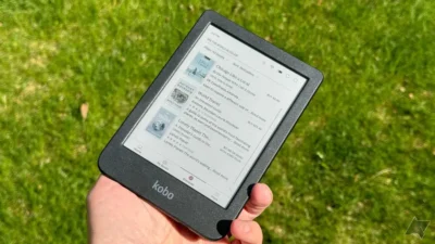 A hand holding a Kobo ereader showing three books listed. The background is blurry green grass.