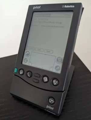 A grey coloured PDA device resting in a cradle on a desk. At the bottom of the PDA are four round buttons, with a small green button to the left, and in the centre bottom are two smaller rectangular buttons for scrolling up or down. Bottom right of the cradle is a single round button.