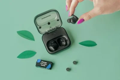 Green background with three green leaves. A small black plastic case rests with its top open. Inside is one black earbud. To the right is another such earbud clasped between a few fingers. To the left is a 9V shaped battery with the words on it: Change is in your hands.