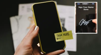 Hand holding up an iPhone with a post-it note stuck on the front that has writing on saying "sign here" with an arrow pointing to the left
