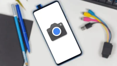 Smartphone camera with a white screen background and just a large icon of a camera showing. The background is blurred but shows a blue pen and pencil to the left, and an adaptor plug to the right with four multi-colour leads extending from it.