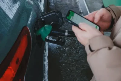 Close view of a petrol pump nozzle inside a car. It is a dark green car and the nozzle is green and black. A person's hands are seen holding a phone. The screen is slightly blurred but shows green and white and looks something like a messenger app.