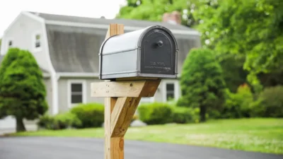 A mailbox sitting on top of a wooden post, with the blurred background showing house with a large green tree to its left.