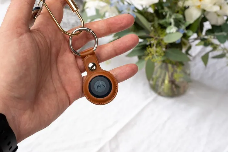 A hand holding a grey and black coloured disc shaped tracker device. The tracker disc is held by a tan coloured leather looking holder, that is attached to a circular silver keyring holder.