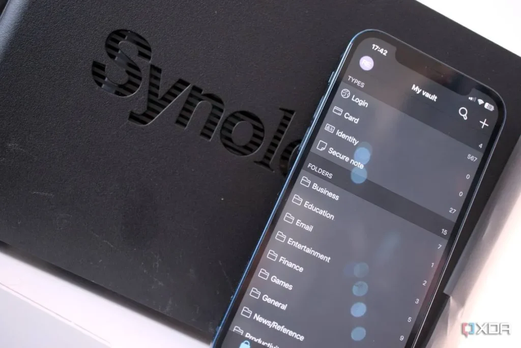 Top of a black plastic cover with cut-out letters showing "Synol". Covering the rest of the word is a smartphone resting on top showing a screen with the title My vault. Down the screen is a list of options such as Login, Card, Identity, Secure notes, Business, Education, Email, etc.