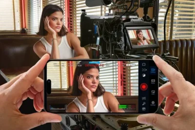 Young woman sitting at a table and facoing a professional grade video camera in front of her. Offset to the left is a pair of hands holdinga smartphone which has the younf woman framed in the display. Overlaid on the view are some HUD controls for shutter speed, ISO, audio level meters, etc.