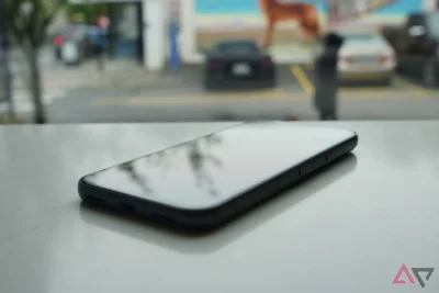 Oblique view of a smartphone resting on a surface. In the background is a blurred view of a street, with two vehicles standing in parking bays.