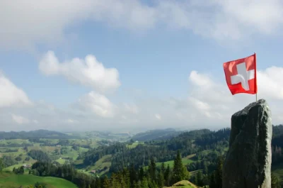 Typical Swiss countryside view with rolling green hills and tall green trees. To the right in the near foreground stands a vertical piece of rock, with a Swiss flag mounted on top of it.