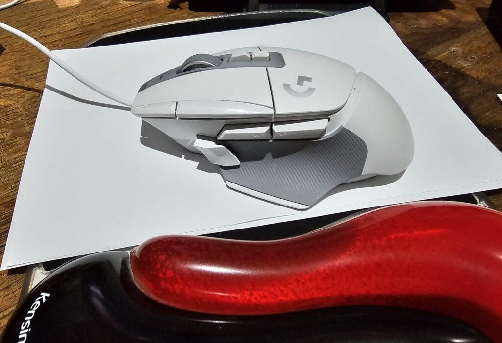 A white coloured gaming mouse with lots of side buttons. It is resting on a piece of white paper. The white paper is on a mouse pad that has a wrist rest in the foreground, which is a red and black colour.