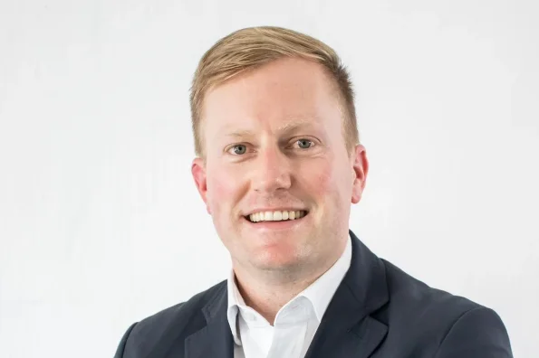 White man with short hair brushed to the side, wearing an open necked white shirt with a collar, and a charcoal coloured jacket.