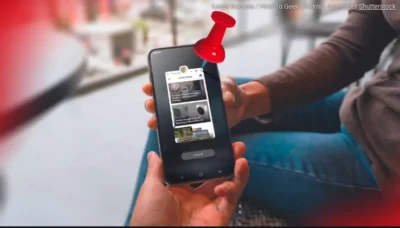 One person's hand, handing a phone to another person. On the phone's screen can be seen an app window with a large red plastic 3D pin stuck into the app.