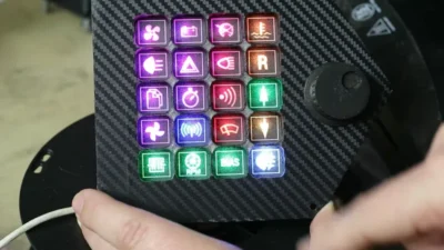 Black box with a round black know that looks like a volume knob. On top of the box can be seen 20 coloured buttons in a grid of 4 wide and 5 high. The buttons have logos on that depic a fan, a headlight, warning triangle, battery, temperature gauge, etc.