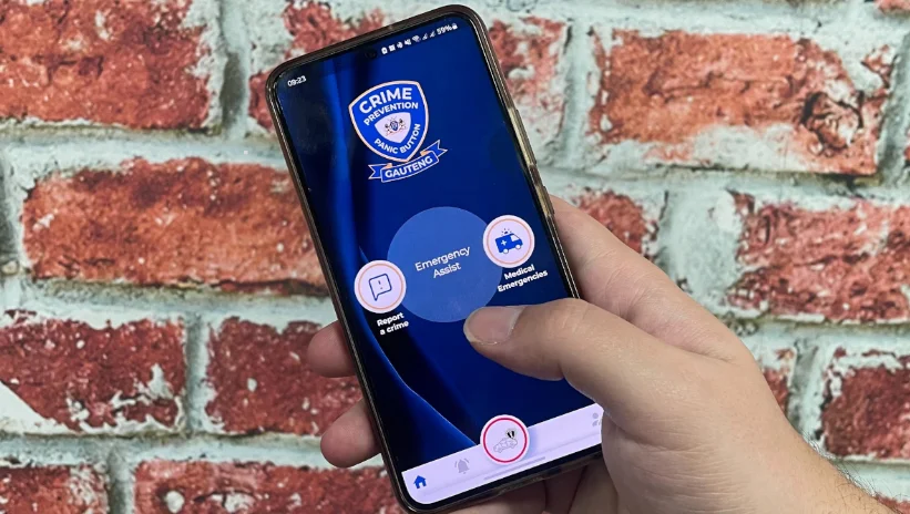 Brick wall background with a hand holding a smartphone. On the screen is a Crime Prevention badge with a big blue button in the centre marked Emergency Assist.