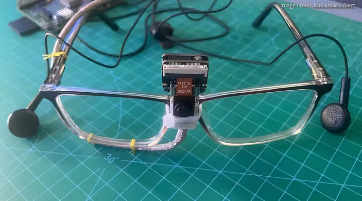 Pair of spectacles resting on a green cutting board. In the centre on top is mounted a small camera sensor with a 2cm square circuit board on the back of it. An earphone piece straddles each arm of the spectacles. In the background is a blurry view of a Raspberry Pi computer where the cables lead to.